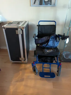 Discover My Mobility blue motorized wheelchair with joystick control, shown alongside a heavy-duty black flight case with aluminum trim.