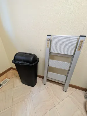 Delxo three-step folding aluminum step stool alongside a black plastic swing-top trash can.
