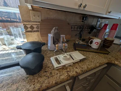 Assorted kitchenware including a mandoline slicer, a spiralizer, two black saucepans with lids, a wooden cutting board, and a Pyxis mug...