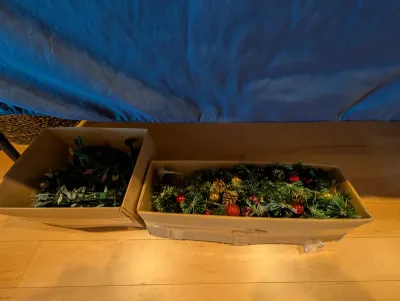 Two cardboard boxes containing Christmas decorations, including a decorated artificial pine garland and a bundle of multi-colored string...