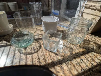 Assorted glass and ceramic vessels, including a hobnail jar, a ribbed green glass bowl, and a square glass holder, displayed on a granite...