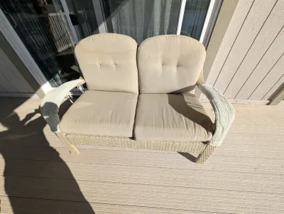 Woven wicker outdoor loveseat with beige cushions, showing some water staining and wear on the armrests.