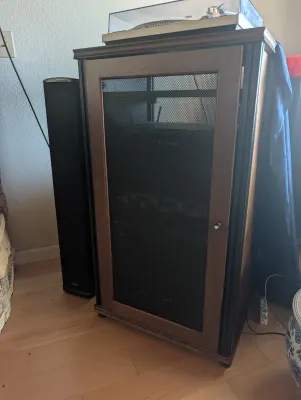 Wooden media cabinet with a mesh front door, housing audio equipment and supporting a turntable on top, flanked by a tall black speaker.