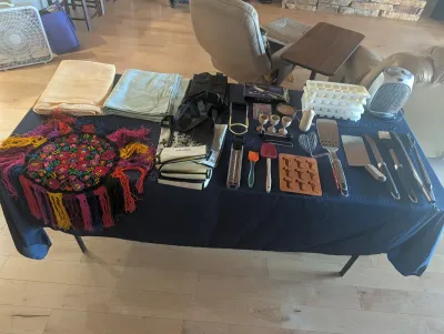 Assorted household items displayed on a blue tablecloth, including folded linens, reusable shopping bags, a colorful fringed textile,...