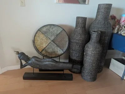 Assorted home decor items including a reclining Buddha statue on a stand, a large decorative metal charger plate, and three tall woven...