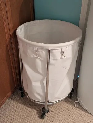 Rolling laundry hamper featuring a white canvas bag suspended on a chrome-finished metal frame with caster wheels.