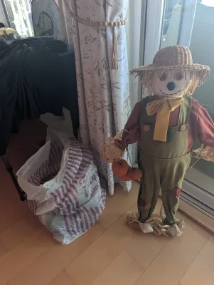 Decorative autumn scarecrow figure wearing green overalls, a red shirt, and a straw hat, standing next to a plastic shopping bag.