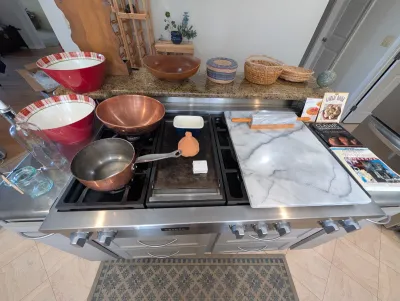 Assorted kitchenware including copper mixing bowls, a marble pastry board with rolling pin, cookbooks, and decorative woven baskets...
