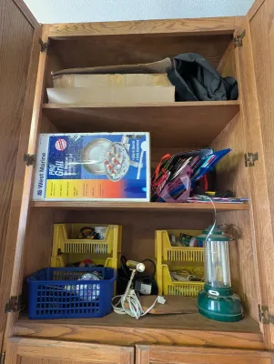 Assorted household and outdoor items stored in a wooden cabinet, including a West Marine Pro Grill in its original box, a green camping...