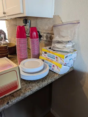 Assortment of disposable party supplies, including stacks of red plastic cups, white plastic plates, and boxes of plastic cutlery.