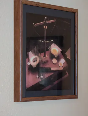 Framed still life photograph depicting an antique balance scale, presented in a dark mat and a simple oak wood frame.