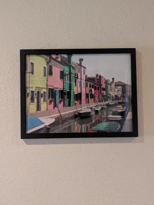 Framed color photograph depicting a canal in Burano, Italy, with vibrant houses and moored boats, housed in a simple black frame.