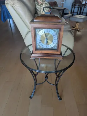 Vintage wooden bracket clock with a brass carrying handle and ornate face, displayed on a small round glass-top side table with a black...