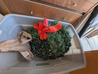Artificial Christmas wreath with a red velvet bow, stored in a grey plastic bin alongside multi-colored string lights.