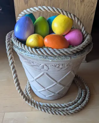 Large decorative Easter basket planter filled with oversized, brightly painted wooden or plaster eggs, featuring a thick rope handle and...
