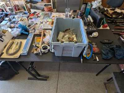 Assorted plumbing and hardware supplies, including a large blue plastic bin filled with various fittings, alongside hand tools, hoses,...