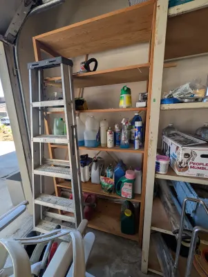 An aluminum stepladder stands in front of a wooden shelving unit filled with various household and automotive chemicals, including...