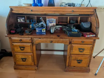 Vintage oak roll-top desk with multiple drawers, cubby compartments, and brass hardware, shown holding various electronics and accessories.
