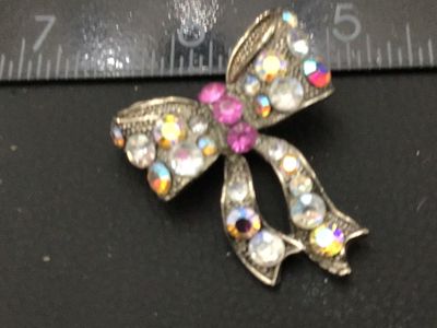 Sale Photo Thumbnail #275: A silver-tone ribbon bow brooch featuring iridescent/aurora borealis and clear rhinestones with pink rhinestone accent stones at the center knot. Measures approximately 1.25 inches across. Features a roll-over safety pin clasp on the back. Good pre-owned 