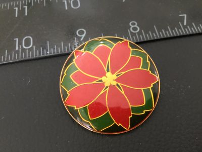 Sale Photo Thumbnail #16: Round enamel brooch featuring a red and green holiday poinsettia design with gold-tone outlines. Measures approximately 1.5 inches in diameter. Shown in good pre-owned condition with smooth enamel finish.