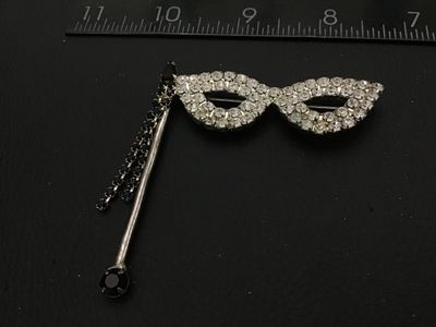 Sale Photo Thumbnail #126: Vintage silver-tone rhinestone brooch shaped like a masquerade opera mask on a stick. Features clear set rhinestones across the mask frame with black rhinestone accents along the handle and dangling fringe details. Secure pin back fastener. Shows light vi