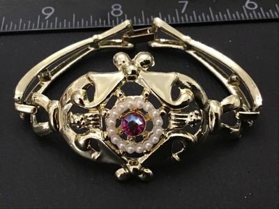 Sale Photo Thumbnail #58: Ornate vintage gold-tone cuff-style bracelet featuring an ornate filigree center medallion set with a halo of faux pearls and a central pink/magenta Aurora Borealis rhinestone. Features a fold-over clasp closure. Measures approximately 7 inches in circumf