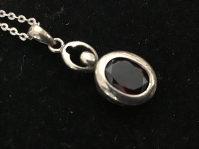 Sale Photo Thumbnail #161: A sterling silver chain necklace featuring an oval-cut faceted dark red stone (possibly garnet) pendant with a small accent stone setting above it. The pendant loop is marked 'LUC' and features textured dot details along the side setting. Includes a delic