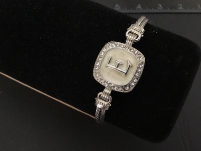 Sale Photo Thumbnail #29: Silver-tone twisted cable style cuff/bangle bracelet featuring a square center emblem with the initial 'E' set against an off-white enamel background, surrounded by a halo of clear rhinestones/crystals. Features a magnetic closure at the back. Light overa
