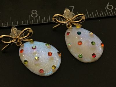 Sale Photo Thumbnail #281: Pair of festive  egg dangle earrings featuring iridescent white glittery teardrop/egg-shaped bases accented with multicolored rhinestones. Topped with gold-tone ribbon bows and post backs with plastic bullet clutch stoppers. Measures approximately 1.5 inc