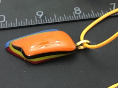 Sale Photo Thumbnail #61: Artisan handcrafted fused glass pendant featuring layered geometric colors including orange, yellow, blue, black, and matte burgundy. Attached with a gold-tone bail to a yellow cord necklace. In good condition with no visible chips or cracks. Measures app