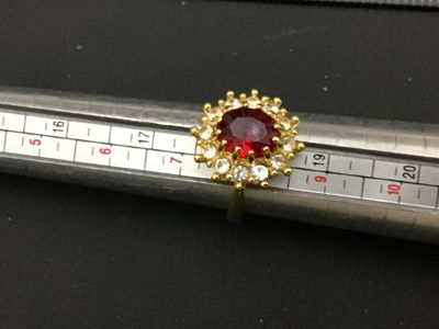 Sale Photo Thumbnail #273: Gold-tone costume jewelry cocktail ring featuring a prominent oval red central stone surrounded by a halo of clear rhinestone accents. Ring measures approximately size 8 on a ring mandrel. Good condition with minimal wear visible.