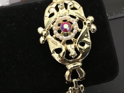 Sale Photo Thumbnail #59: Ornate vintage gold-tone cuff-style bracelet featuring an ornate filigree center medallion set with a halo of faux pearls and a central pink/magenta Aurora Borealis rhinestone. Features a fold-over clasp closure. Measures approximately 7 inches in circumf