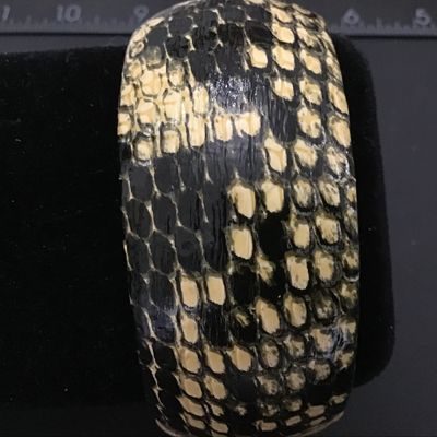 Sale Photo Thumbnail #92: A wide, chunky bangle bracelet featuring a black and cream python/snakeskin pattern finish over a hard core. The interior is lined in black and retains a remnant of an old price tag. Shows light vintage wear consistent with age.