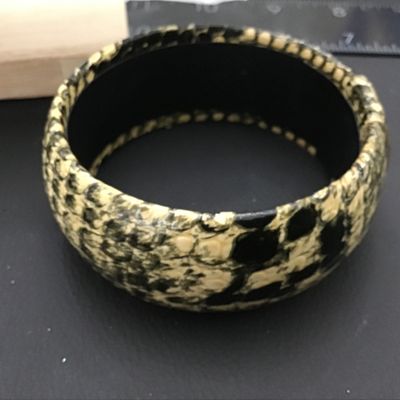 Sale Photo Thumbnail #94: A wide, chunky bangle bracelet featuring a black and cream python/snakeskin pattern finish over a hard core. The interior is lined in black and retains a remnant of an old price tag. Shows light vintage wear consistent with age.