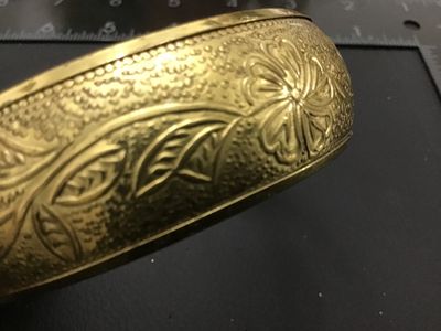 Sale Photo Thumbnail #218: A vintage gold-tone brass bangle bracelet featuring an intricate embossed floral and leaf design with textured background details. Inner diameter measures approximately 2.5 inches across. Shows minor age-appropriate wear and light patina.