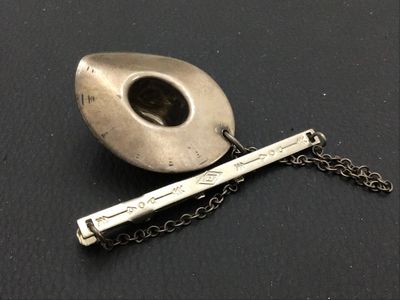 Sale Photo Thumbnail #252: Vintage Western-style tie bar clip featuring a detailed cowboy hat charm attached via safety chain. The tie bar features stamped arrow and geometric motifs on the front. Silver metal shows aged patina and light surface wear consistent with age. Marked ste
