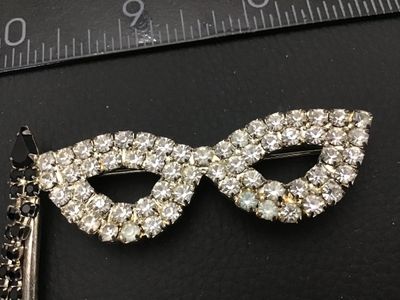 Sale Photo Thumbnail #127: Vintage silver-tone rhinestone brooch shaped like a masquerade opera mask on a stick. Features clear set rhinestones across the mask frame with black rhinestone accents along the handle and dangling fringe details. Secure pin back fastener. Shows light vi