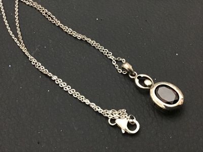 Sale Photo Thumbnail #163: A sterling silver chain necklace featuring an oval-cut faceted dark red stone (possibly garnet) pendant with a small accent stone setting above it. The pendant loop is marked 'LUC' and features textured dot details along the side setting. Includes a delic