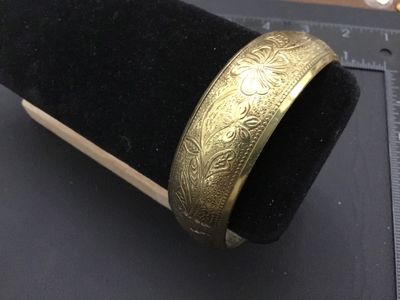 Sale Photo Thumbnail #220: A vintage gold-tone brass bangle bracelet featuring an intricate embossed floral and leaf design with textured background details. Inner diameter measures approximately 2.5 inches across. Shows minor age-appropriate wear and light patina.