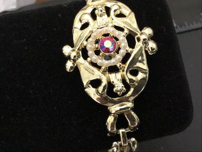 Sale Photo Thumbnail #57: Ornate vintage gold-tone cuff-style bracelet featuring an ornate filigree center medallion set with a halo of faux pearls and a central pink/magenta Aurora Borealis rhinestone. Features a fold-over clasp closure. Measures approximately 7 inches in circumf