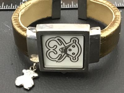 Sale Photo Thumbnail #239: TOUS ladies quartz wrist watch featuring a square face with the iconic TOUS bear graphic on the dial and dangling bear charm. Features a stainless steel back, Japan quartz movement, and an open cuff-style band with textured bronze/gold tone finish. Light 