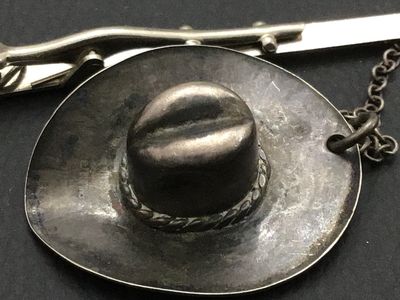 Sale Photo Thumbnail #250: Vintage Western-style tie bar clip featuring a detailed cowboy hat charm attached via safety chain. The tie bar features stamped arrow and geometric motifs on the front. Silver metal shows aged patina and light surface wear consistent with age. Marked ste