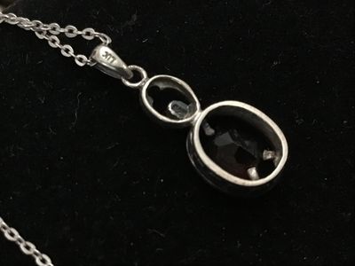 Sale Photo Thumbnail #162: A sterling silver chain necklace featuring an oval-cut faceted dark red stone (possibly garnet) pendant with a small accent stone setting above it. The pendant loop is marked 'LUC' and features textured dot details along the side setting. Includes a delic