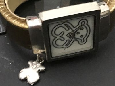 Sale Photo Thumbnail #237: TOUS ladies quartz wrist watch featuring a square face with the iconic TOUS bear graphic on the dial and dangling bear charm. Features a stainless steel back, Japan quartz movement, and an open cuff-style band with textured bronze/gold tone finish. Light 