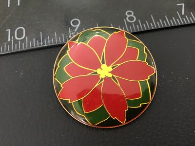Sale Photo Thumbnail #15: Round enamel brooch featuring a red and green holiday poinsettia design with gold-tone outlines. Measures approximately 1.5 inches in diameter. Shown in good pre-owned condition with smooth enamel finish.