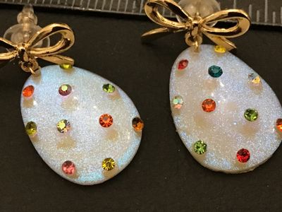 Sale Photo Thumbnail #280: Pair of festive  egg dangle earrings featuring iridescent white glittery teardrop/egg-shaped bases accented with multicolored rhinestones. Topped with gold-tone ribbon bows and post backs with plastic bullet clutch stoppers. Measures approximately 1.5 inc