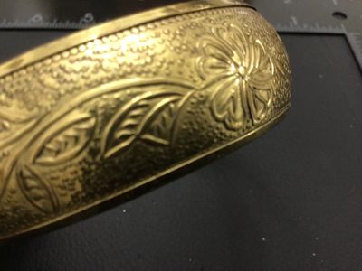 Sale Photo Thumbnail #221: A vintage gold-tone brass bangle bracelet featuring an intricate embossed floral and leaf design with textured background details. Inner diameter measures approximately 2.5 inches across. Shows minor age-appropriate wear and light patina.