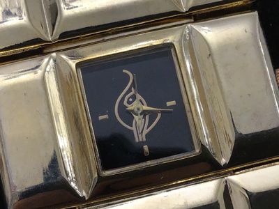 Sale Photo Thumbnail #232: A vintage gold-tone fashion watch featuring a wide, geometric segmented link band and a small square dark dial with stylized graphic logo and minimalist hour markers. Measures Workingapproximately 7.5 to 8 inches in total length. Stainless steel or metal 