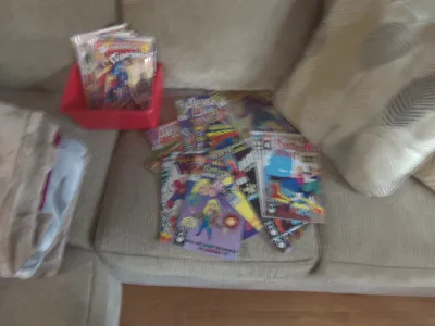 Sale Photo Thumbnail #6: A collection of assorted vintage and modern comic books, featuring titles like Superman, Fantastic Four, and Avengers, displayed on a sofa and inside a red plastic bin.