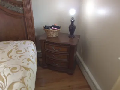 Sale Photo Thumbnail #24: Traditional wooden three-drawer nightstand featuring carved accents and ornate brass bail handles, topped with a dark glass table lamp and woven basket.